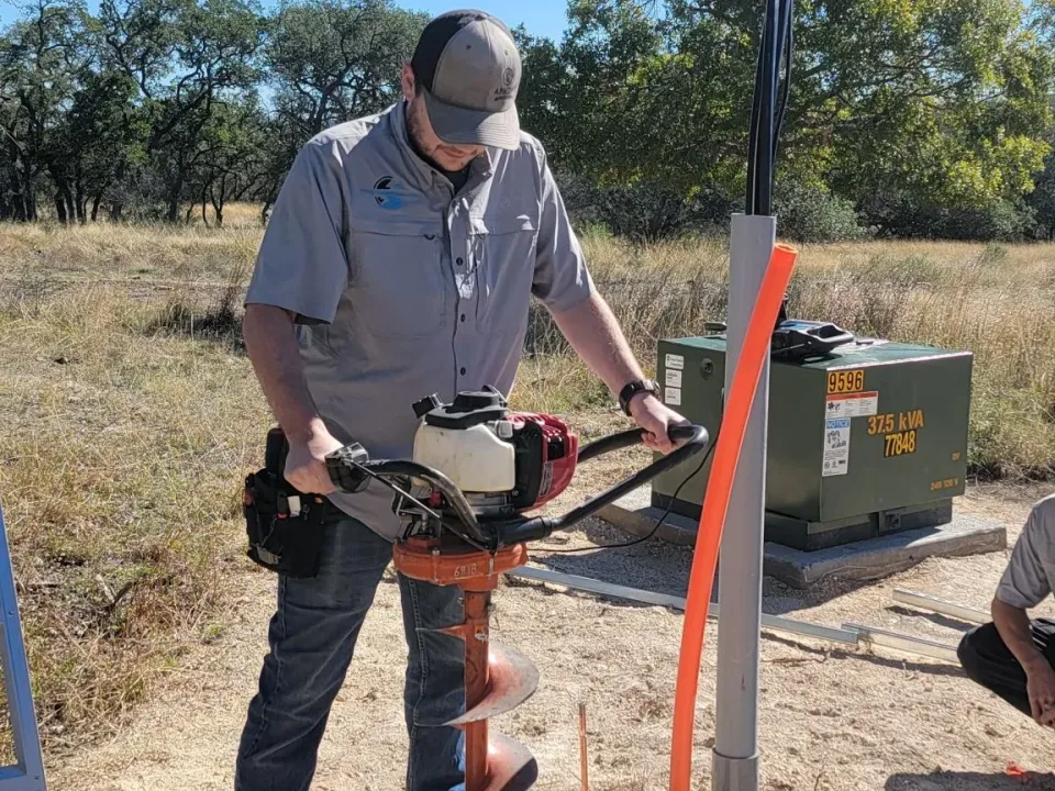 Electrician using a power auger beside a pad-mount transformer with an orange conduit riser